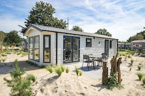 Een kleine, gelijkvloerse hut met een glazen deur en ramen, een terras met een tafel en stoelen, en een zandachtige omgeving.