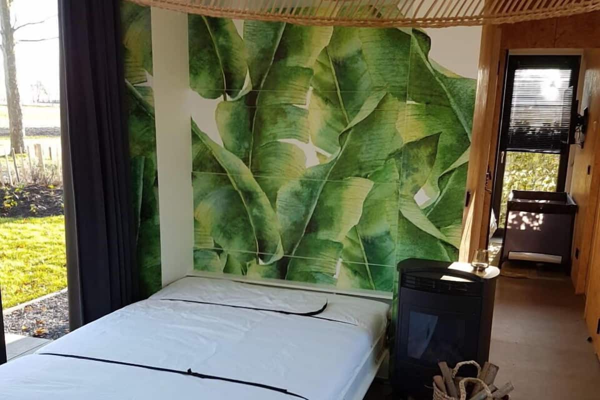 A bedroom with a bed, a wall with a leaf pattern, a small table with a vase and candle, and a basket of logs.