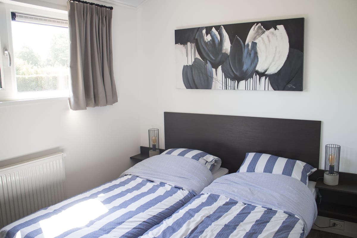 A bedroom with two beds, a window with curtains, a wall-mounted painting, bedside tables with lamps, and a radiator.