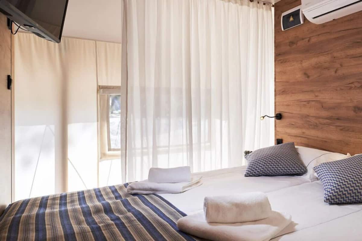 A hotel room with a wooden headboard, a bed with striped bedding, two patterned pillows, a white towel, and a window with sheer curtains.
