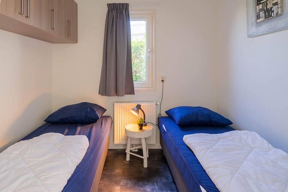A small room with two beds, a window, a radiator, and a desk with a lamp.