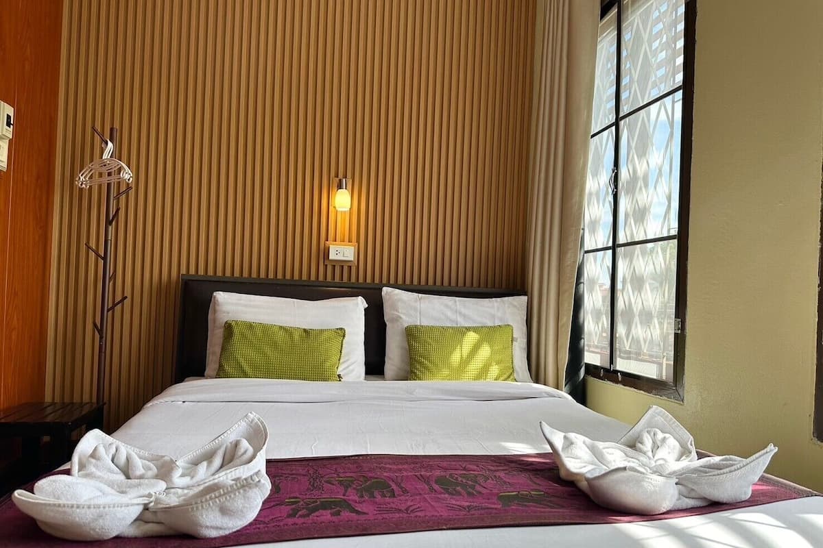 A hotel room with a bed, two pillows, a bedside lamp, a window with a view, and towels on the bed.