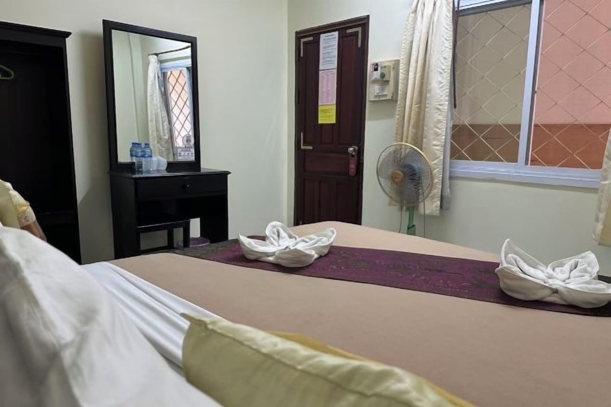 A hotel room with a bed, a dresser, a mirror, a door with a notice, a window with curtains, and a fan.