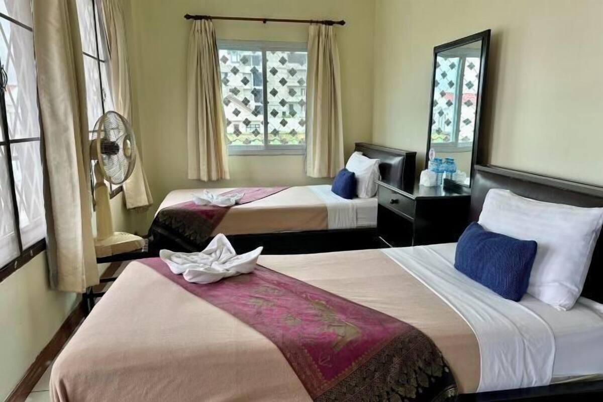 A hotel room with two beds, a window with curtains, a standing fan, a mirror, and a bedside table with toiletries.