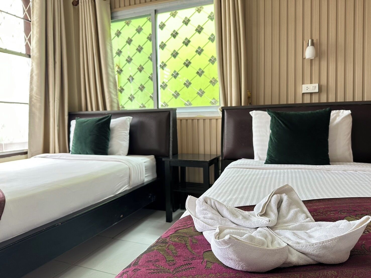 A hotel room with two beds, a window with patterned curtains, a bedside table, and a towel on the floor.