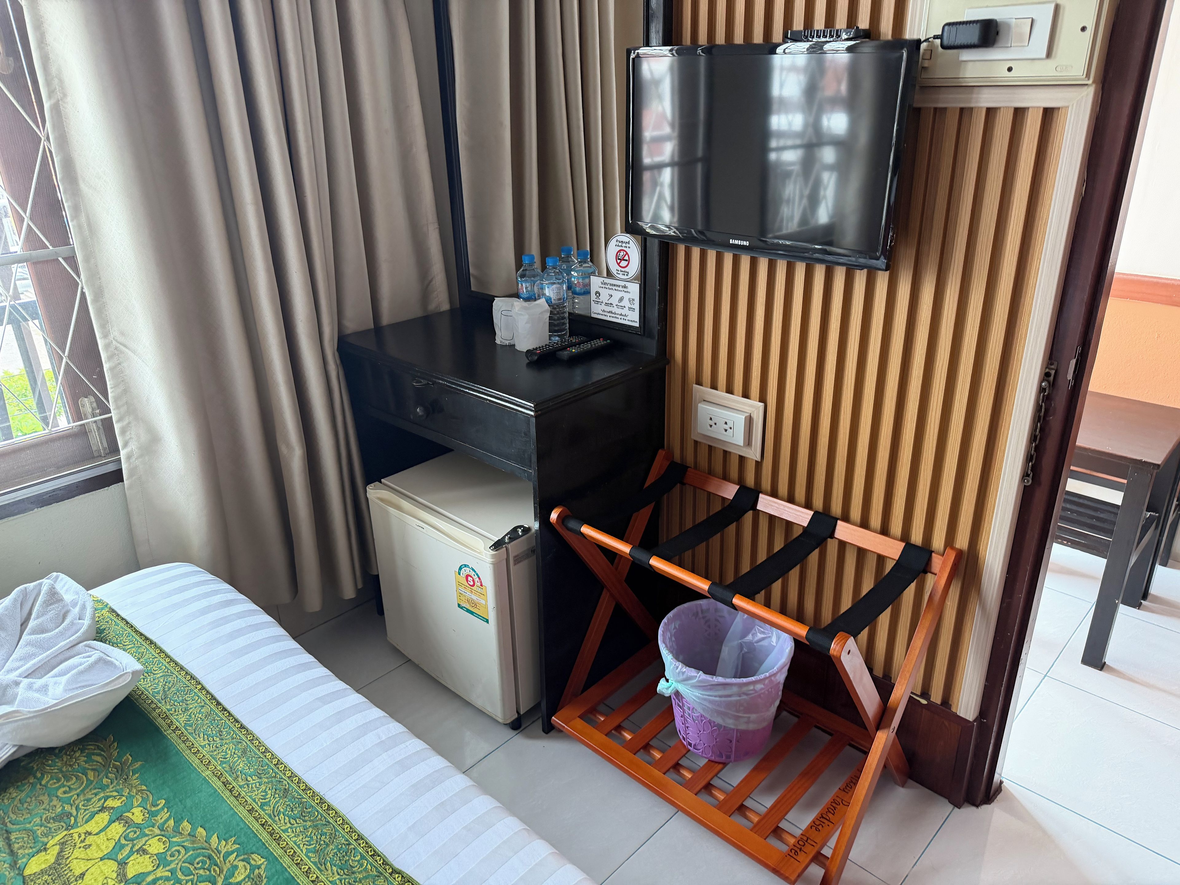A hotel room with a bed, a desk with a television, a small refrigerator, a wooden chair with a tray, and a window with curtains.