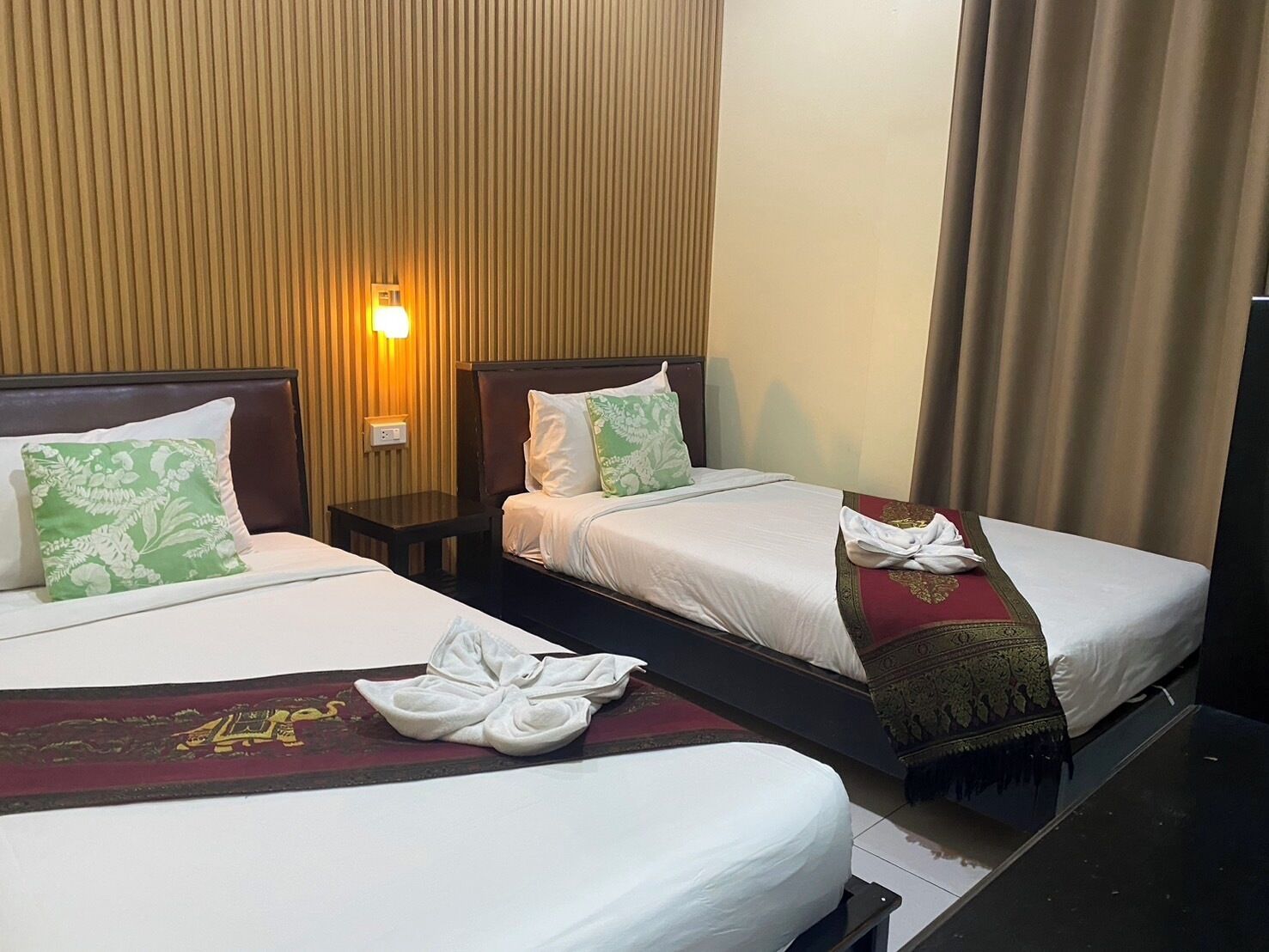 Deux lits côte à côte dans une chambre d’hôtel, avec du linge de lit blanc, des coussins à motifs floraux verts et un chemin de table décoratif rouge.