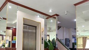 A modern elevator with a metallic finish, a reception desk with a wooden frame, and a large potted plant in the lobby area. - Mixay Paradise Hotel (Vientiane)