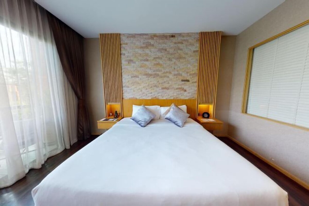 Una habitación de hotel con una cama grande, dos mesitas de noche, un cabecero con un revestimiento de pared texturizado y una ventana con cortinas.