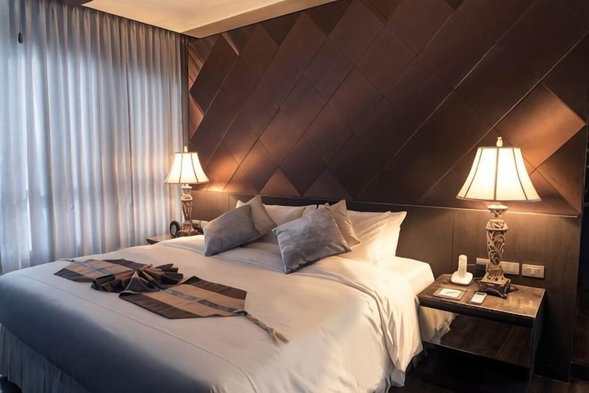 Una habitación de hotel con una cama grande, dos mesitas de noche con lámparas, un cabecero y una cortina.