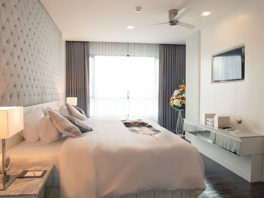 Ein modernes Hotelzimmer mit einem großen Bett, einem Nachttisch mit Lampe, einem an der Wand montierten Flachbildfernseher und einem großen Fenster mit Vorhängen.