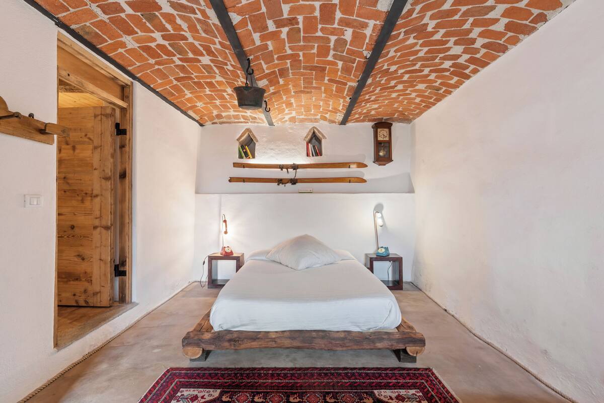 A bedroom with a brick ceiling, wooden bed frame, two bedside tables with lamps, and a red patterned rug.