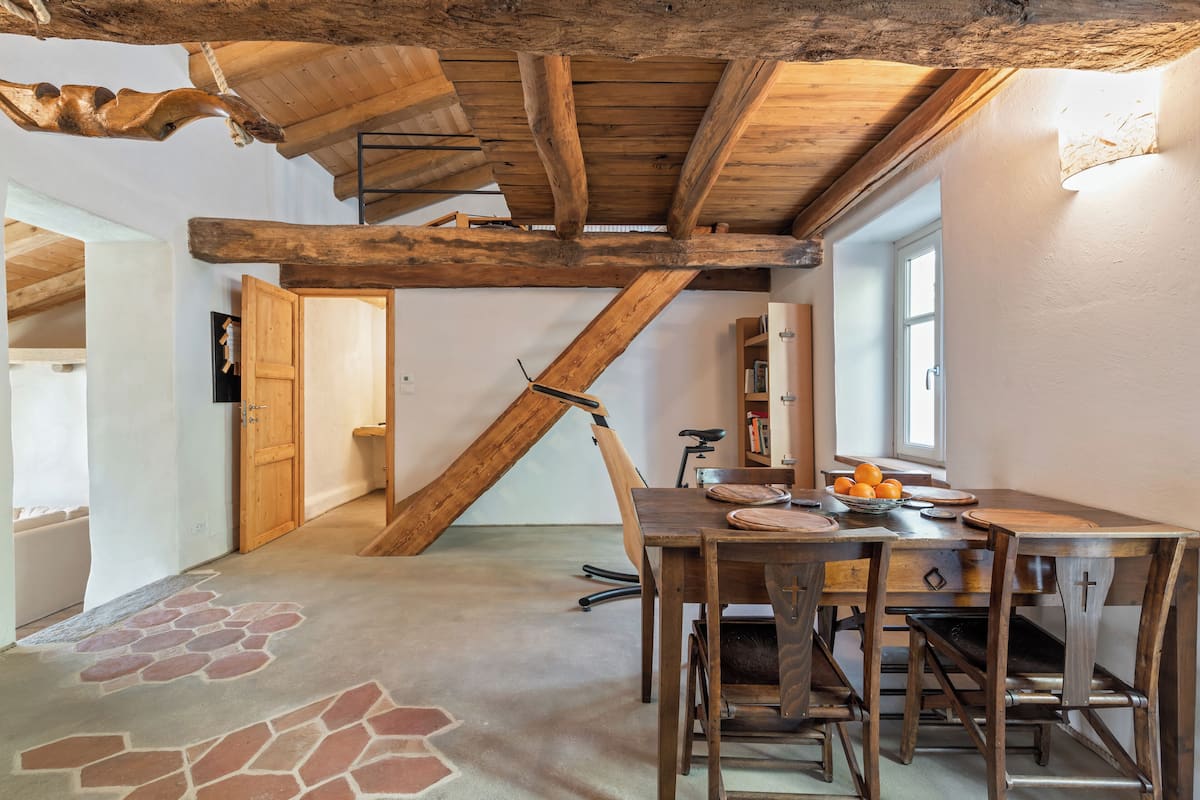 A rustic dining area with exposed wooden beams, a stone-patterned floor, and a wooden table set with plates and oranges.
