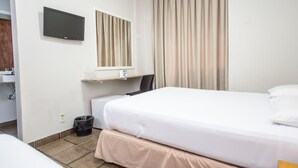 Habitación de hotel con cama, televisor montado en la pared, ventana con cortinas, escritorio con silla y cubo de basura.