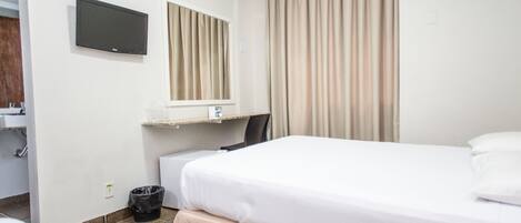 Habitación de hotel con cama, televisor montado en la pared, ventana con cortinas, escritorio con silla y cubo de basura.