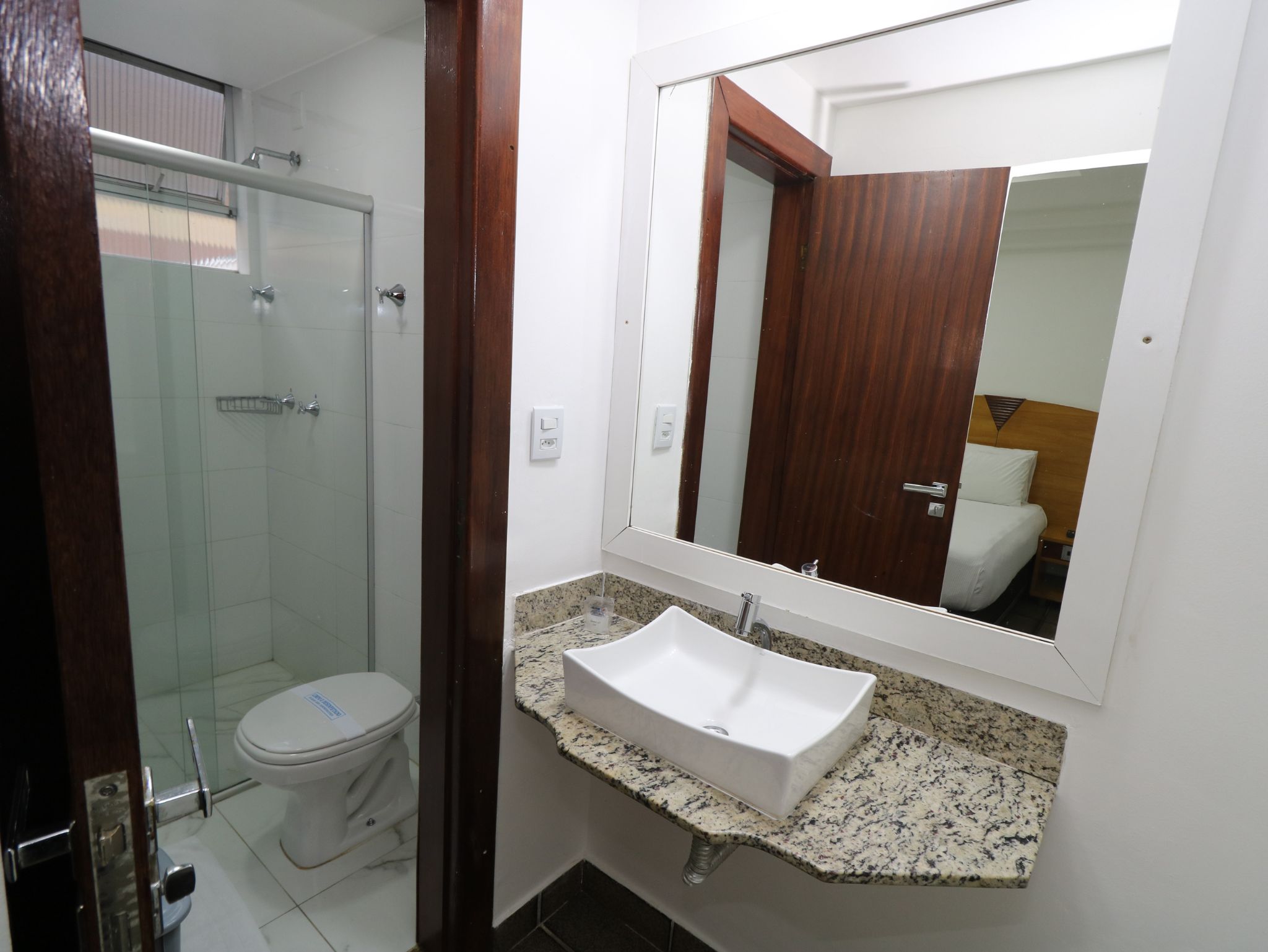 Baño con cabina de ducha de vidrio, inodoro blanco, lavamanos con encimera de granito y espejo con reflejo de puerta de madera.