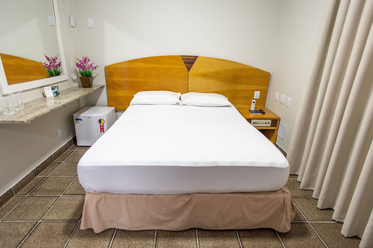 Una cama bien hecha con cabecera, una mesita de noche pequeña con lámpara y un espejo sobre la cama.