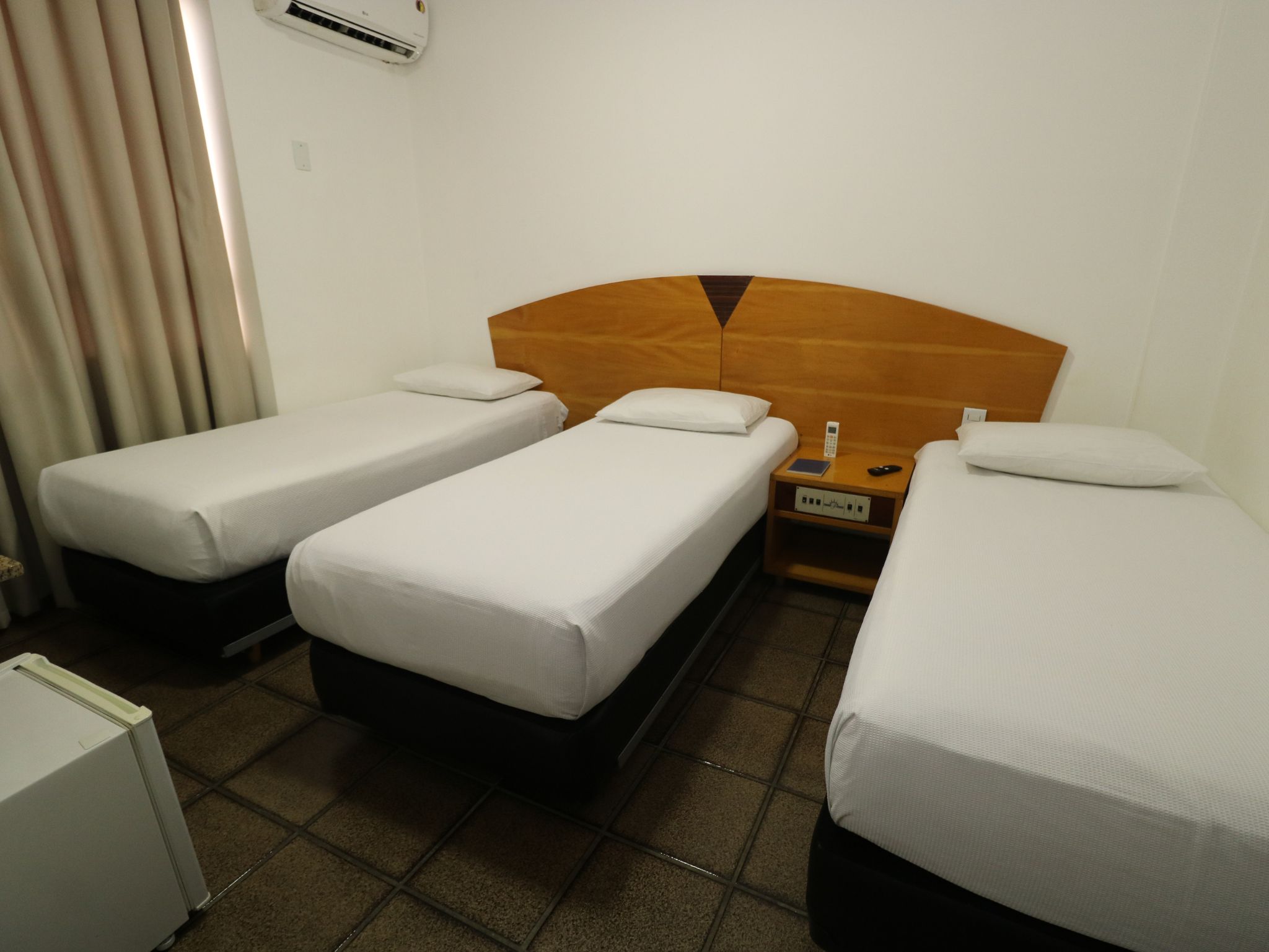 Habitación de hotel con tres camas individuales, una mesita de noche con teléfono y control remoto, y un pequeño armario blanco.