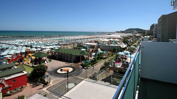 A beachside view with a large number of umbrellas and beach chairs, a playground, and a building with a green roof. - Hotel Excelsior (Cattolica)