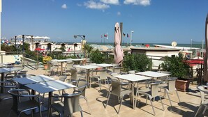 A terrace with tables and chairs, overlooking a beach and the sea. - Hotel Excelsior (Cattolica)