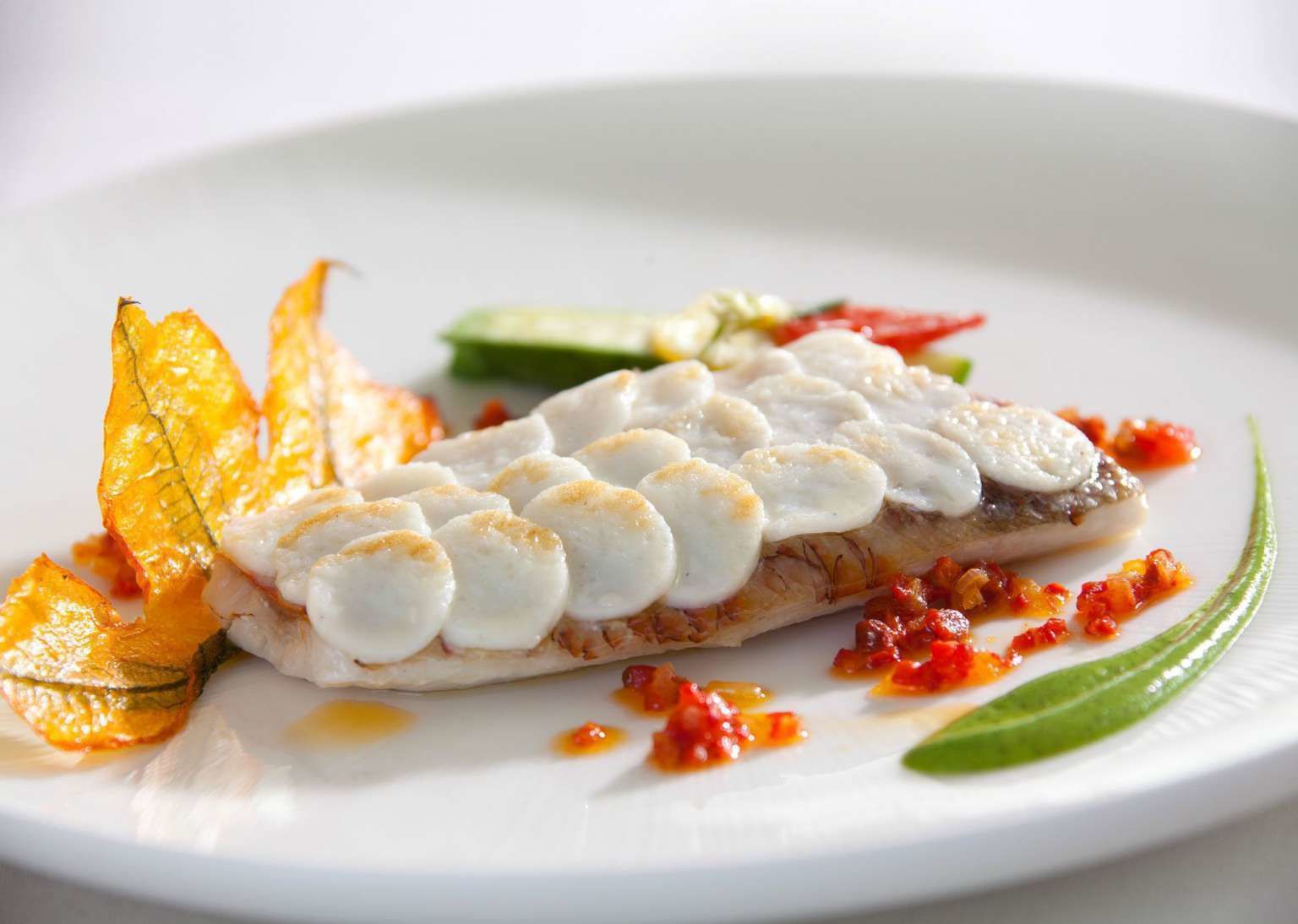 Un plato blanco con un filete de pescado cubierto de láminas blancas, acompañado de verduras y salsa roja.