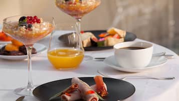Una mesa dispuesta con un vaso de jugo de naranja, una taza de café, un plato con carne enrollada y un postre en un vaso.