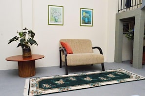 A room with a round wooden table, a beige armchair with a red cushion, a green floral rug, and two framed paintings on the wall. - Pousada Villa Três (Serra Negra)