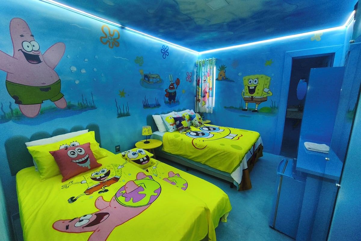 Une chambre avec deux lits, chacun recouvert d’une couette aux motifs de Bob l’éponge, un mur décoré d’une fresque représentant Bob l’éponge et Patrick, et une porte dotée d’un cadre bleu.