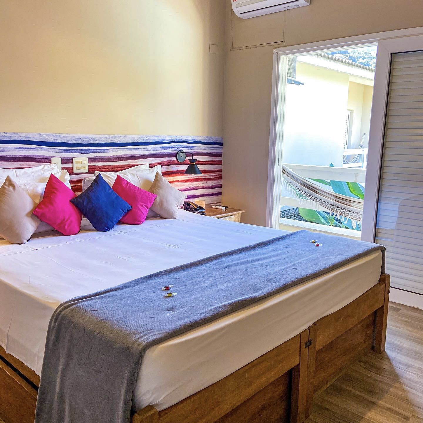 Una cama bien hecha con almohadas coloridas, una lámpara de noche y vistas a un balcón con barandilla.