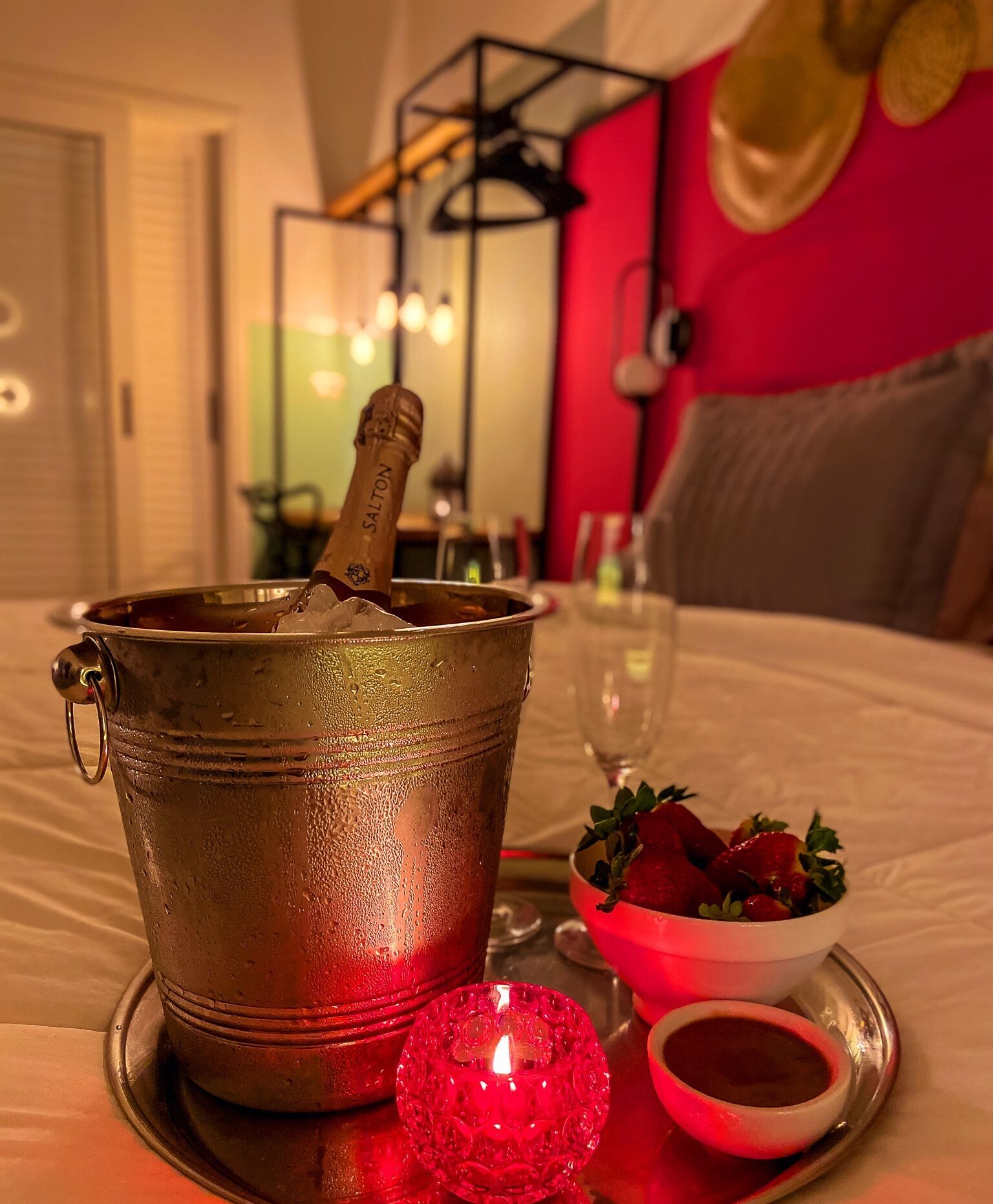Um quarto com uma cama, um balde de metal com uma garrafa, uma tigela de morangos e uma vela sobre uma bandeja.