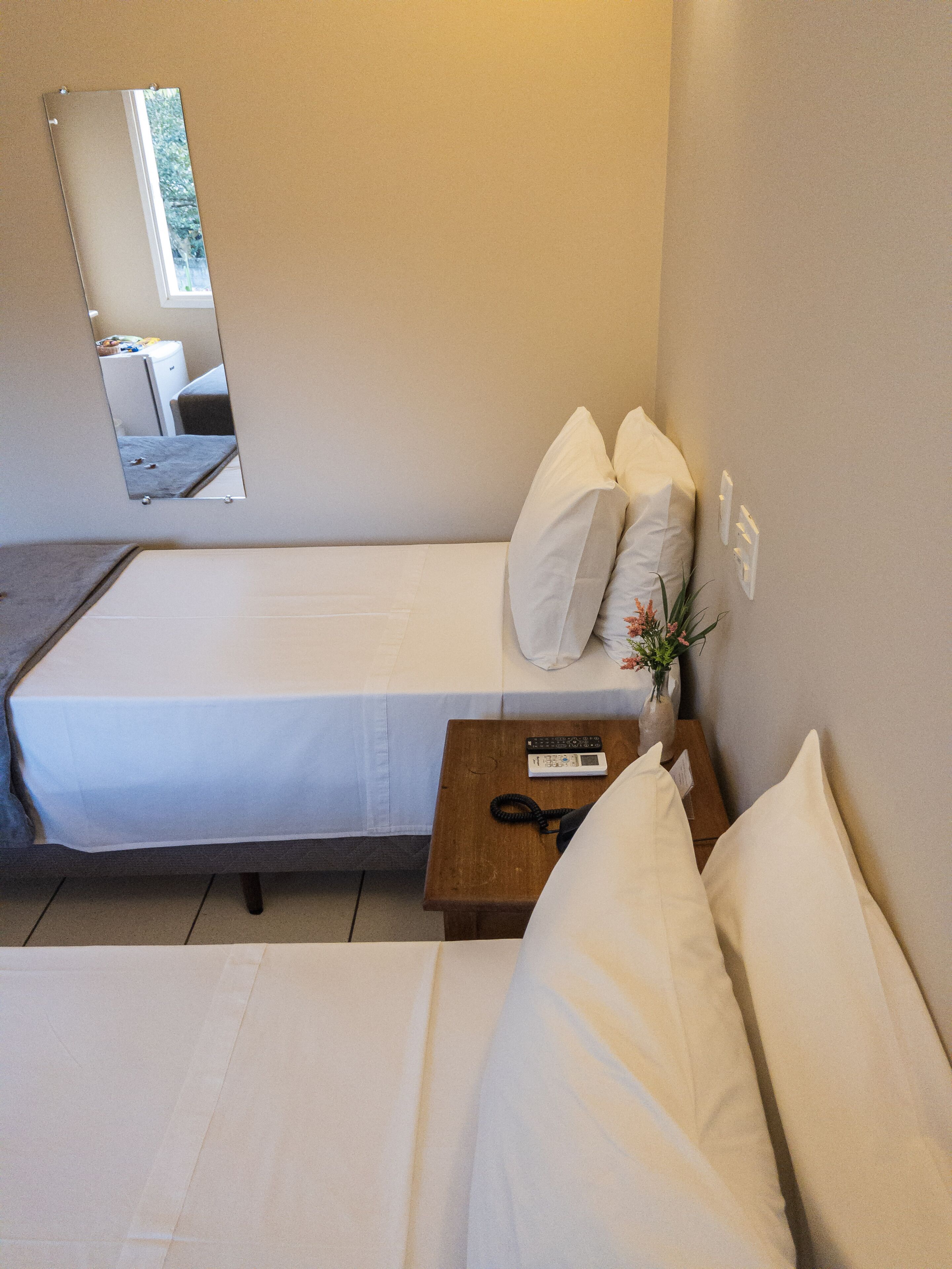 Un lit simple avec une literie blanche, une petite table de chevet avec une télécommande et un miroir qui reflète une pièce avec une fenêtre.