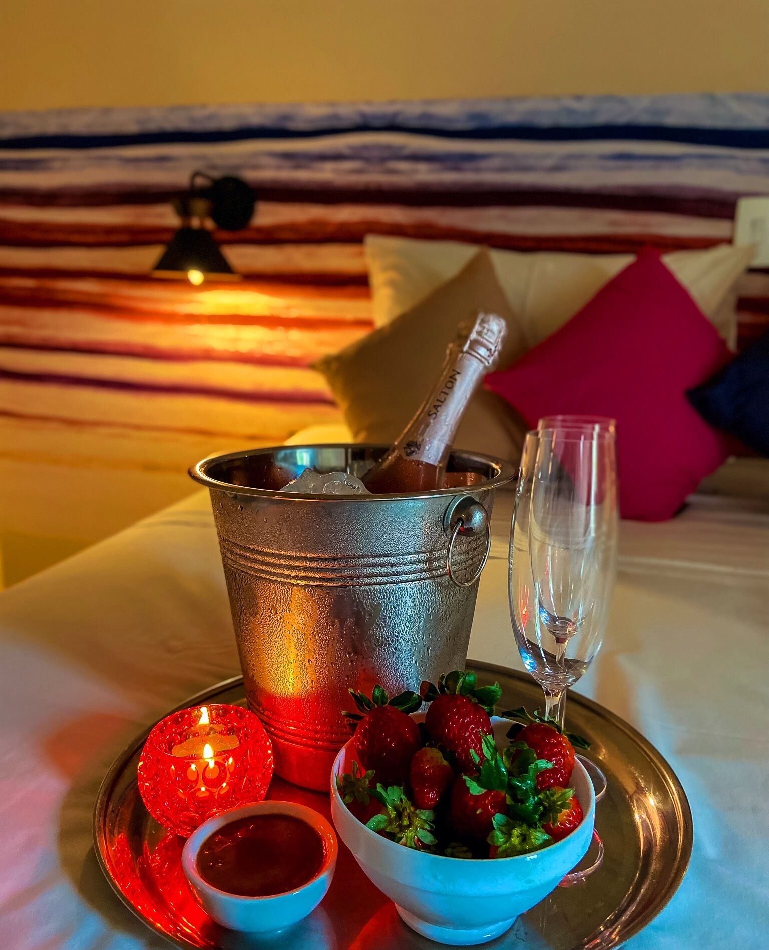 Una cama con un balde de metal que contiene una botella de champán, una copa de champán, un cuenco de fresas y una vela roja sobre una bandeja.