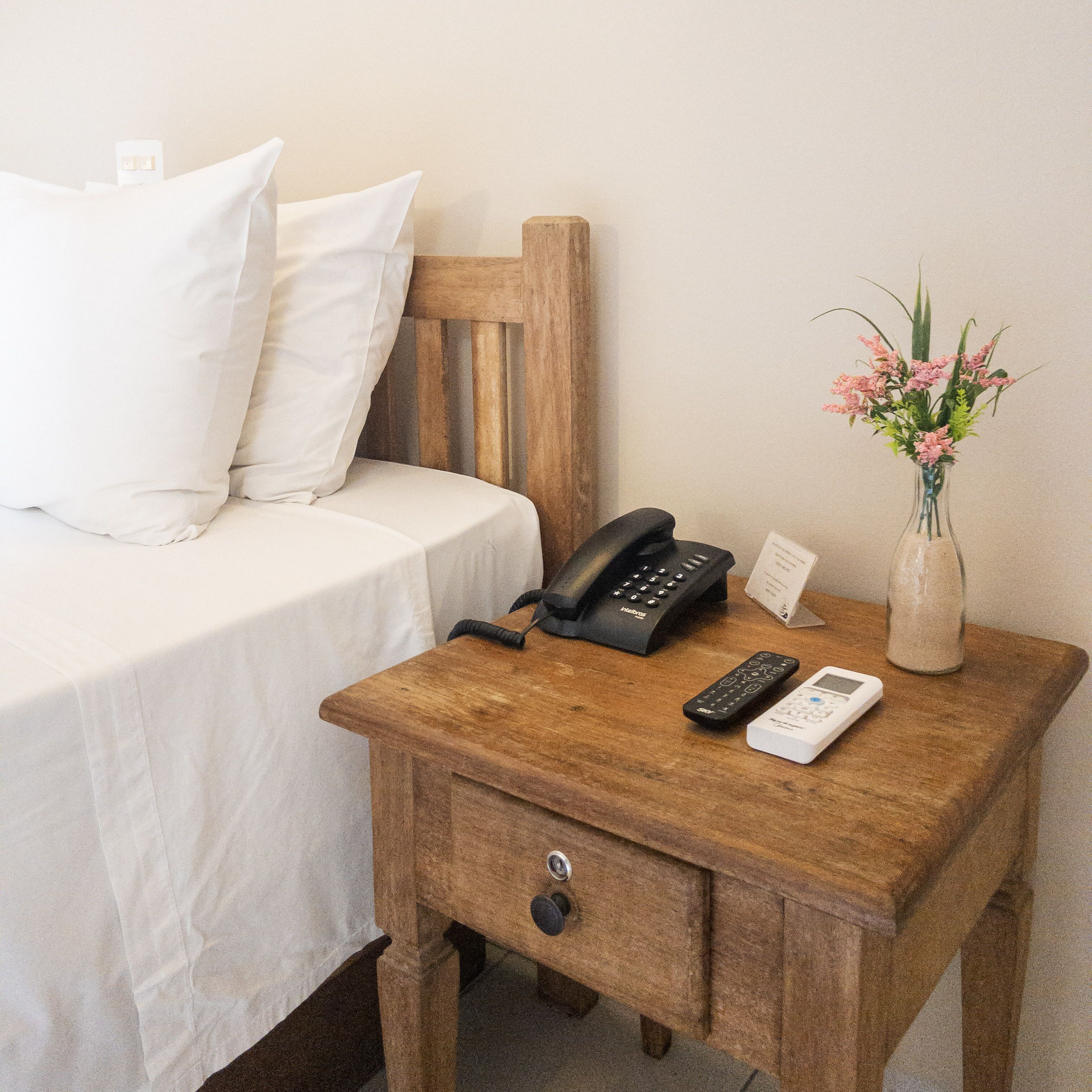 A wooden nightstand with a phone, remote controls, and a vase with flowers.