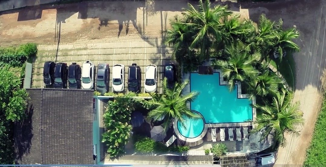 An aerial view of a hotel with a swimming pool surrounded by palm trees, a parking area with several cars, and a covered area with a roof.