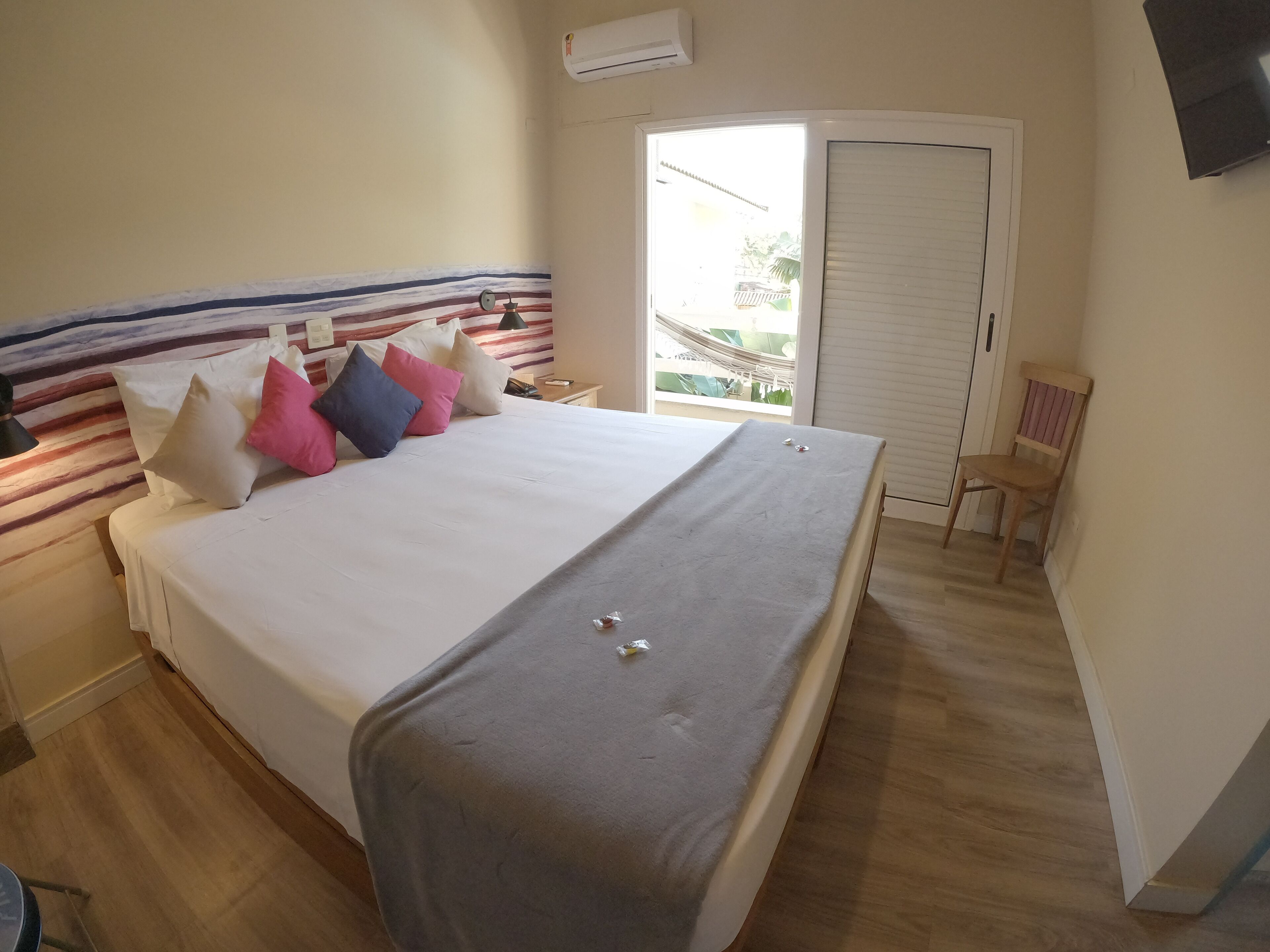 Una cama bien hecha con una colcha gris y varias almohadas de distintos colores. Junto a la cama, una silla de madera y una mesita. Se ve una ventana con persianas, y un equipo de aire acondicionado está montado en la pared.