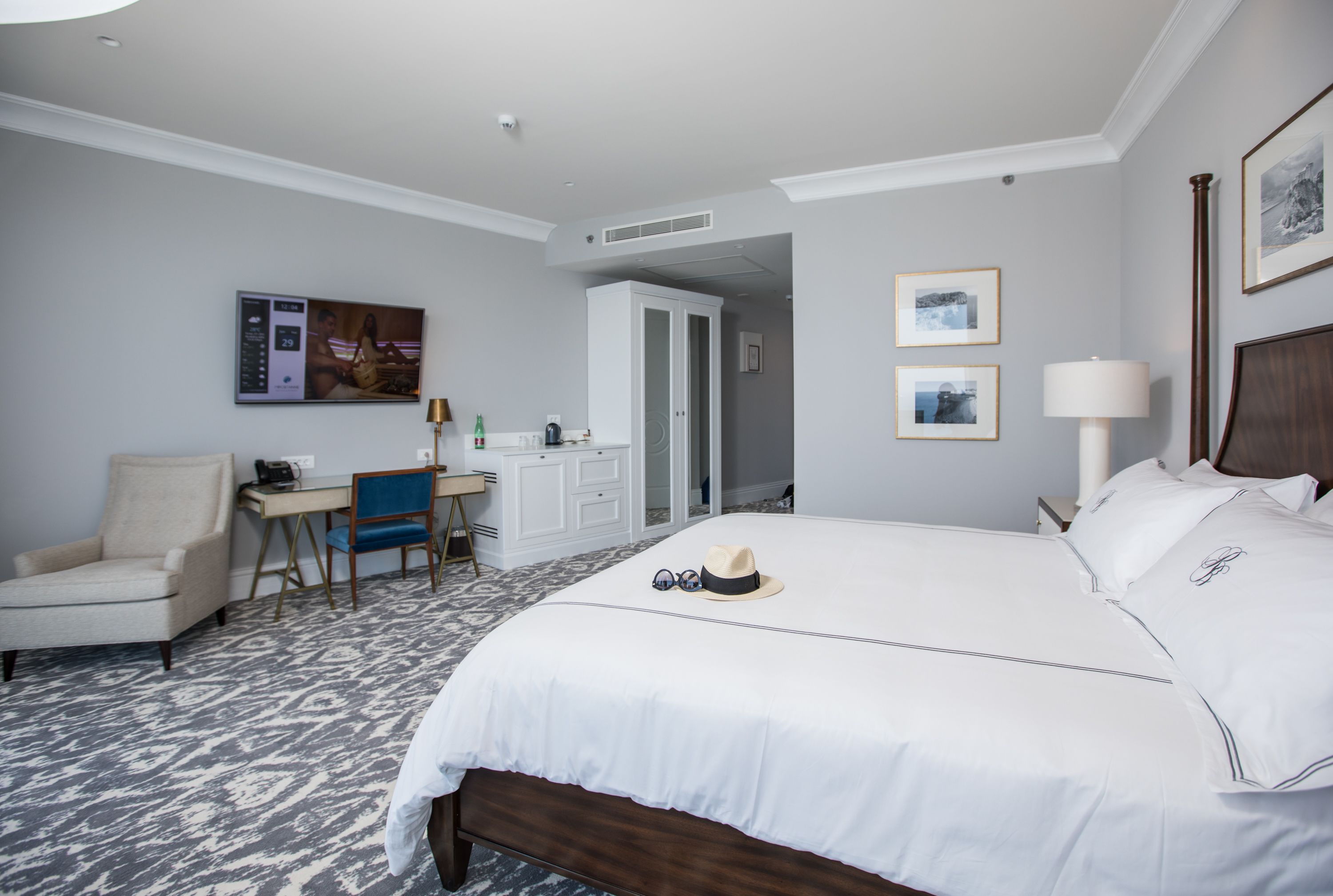 A hotel room with a large bed, a desk with a chair, a TV, and framed pictures on the wall.