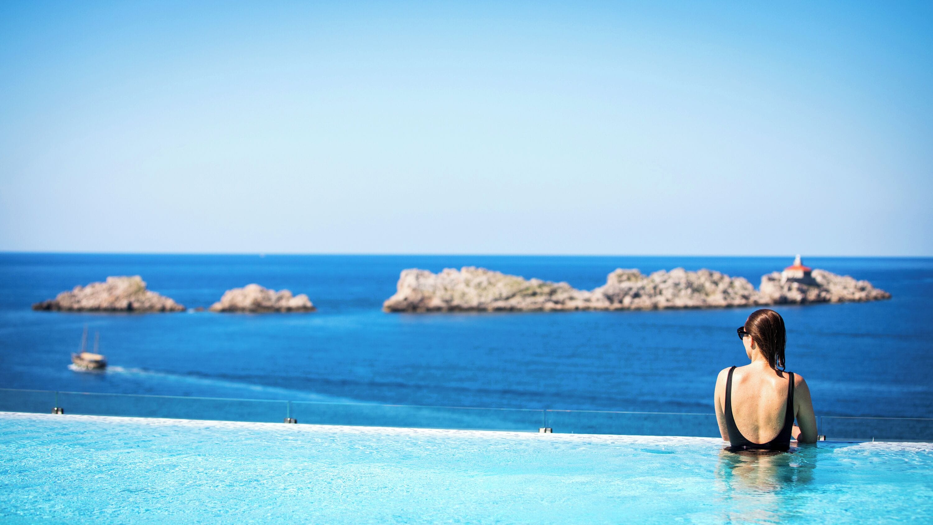 Une femme se trouve dans une piscine qui donne sur une mer bleue et limpide, avec des affleurements rocheux et un phare visible au loin.