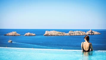 Une femme se trouve dans une piscine qui donne sur une mer bleue et limpide, avec des affleurements rocheux et un phare visible au loin.