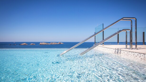 Una piscina azul cristalina con una escalera metálica que da a una terraza con vistas al océano.