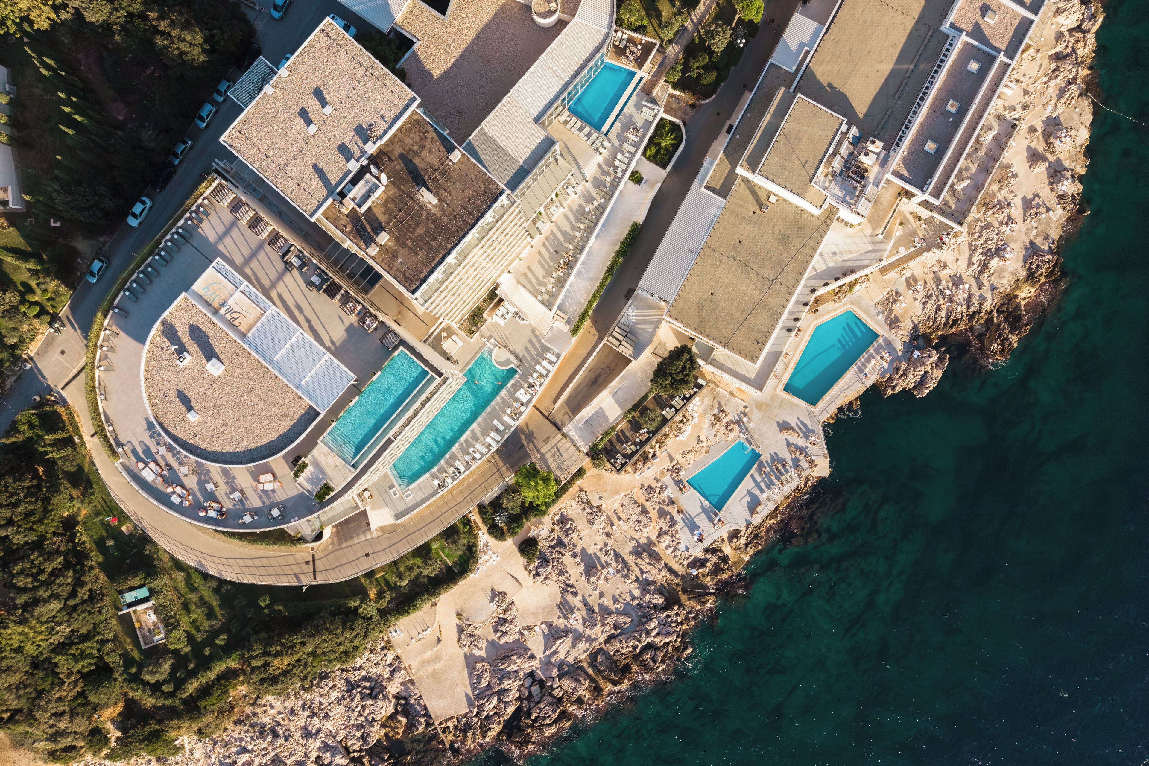 An aerial view of a coastal property with multiple swimming pools, a parking area, and a rocky shoreline.