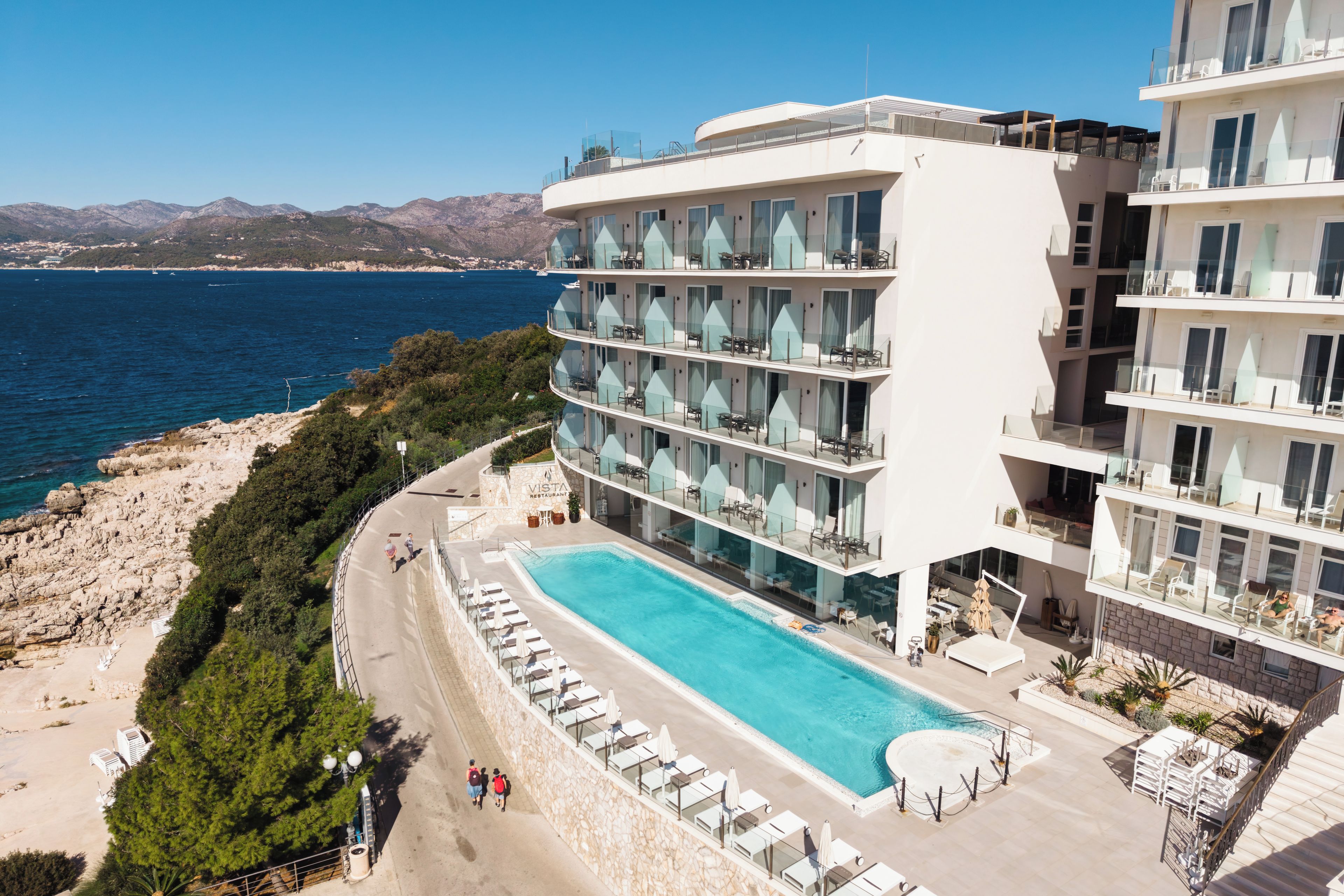 Ein mehrstöckiges Hotel mit Pool, mit Blick auf eine Küstenlandschaft und dahinter Bergen.