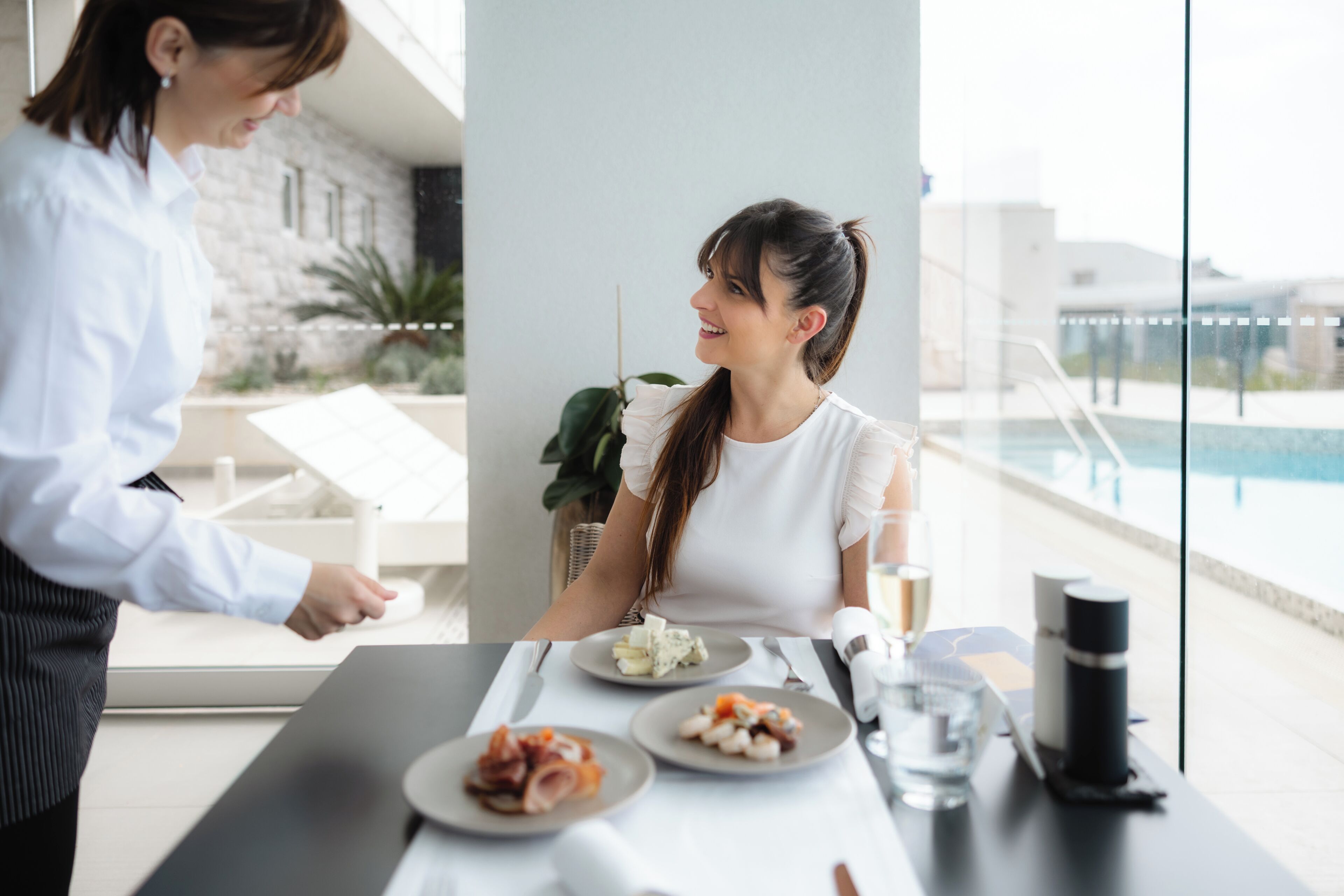 Un espace repas avec vue sur la piscine, un serveur et une table dressée avec des assiettes de nourriture.