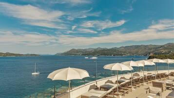 Eine Seeterrasse mit Liegestühlen und Sonnenschirmen, mit Blick auf ein ruhiges Meer mit Booten und einer Bergkulisse.