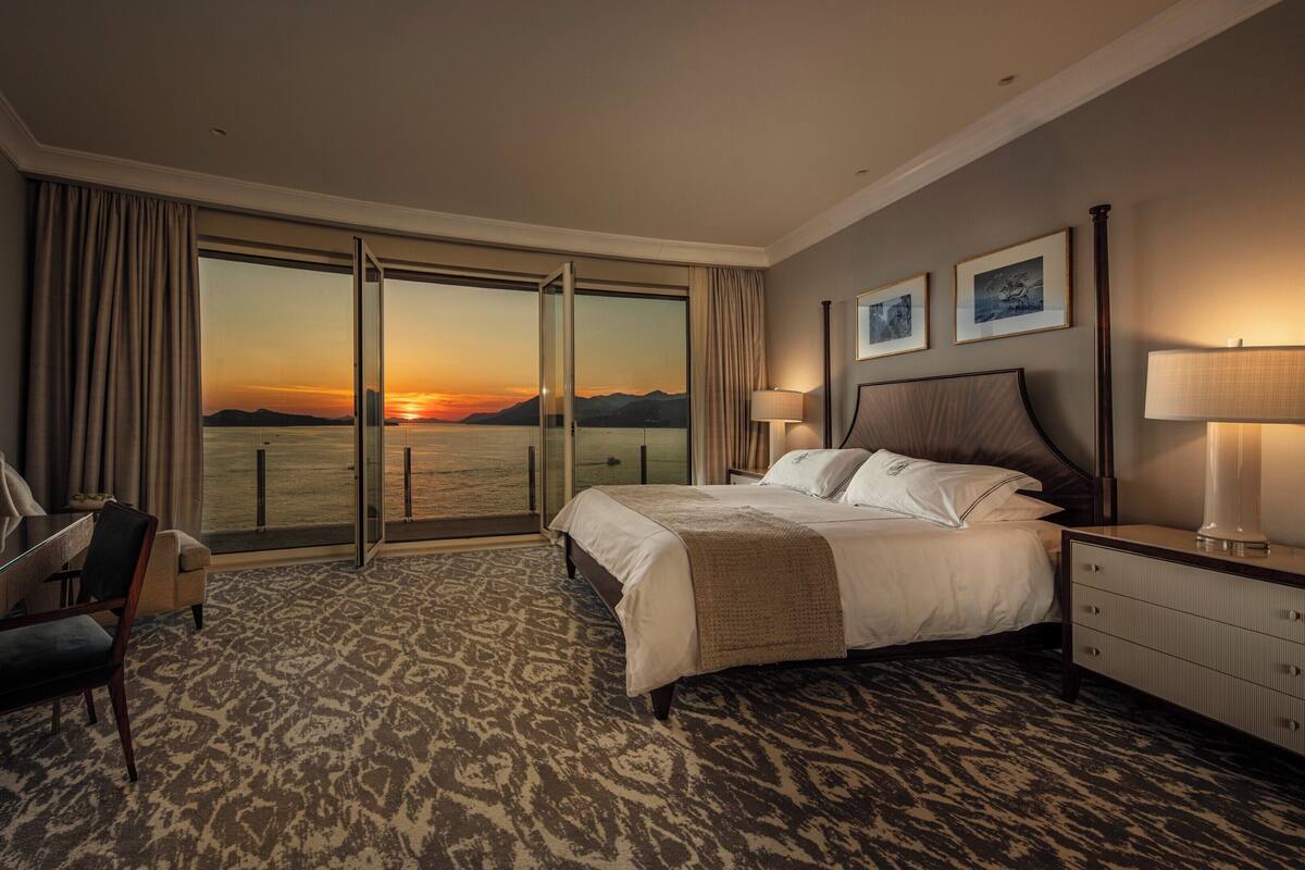Habitación de hotel con cama, escritorio, silla, lámpara y vista al atardecer sobre el agua.