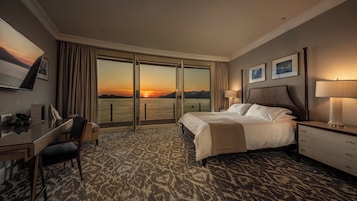 A hotel room with a bed, a desk, a chair, a lamp, and a view of the sunset over the water.