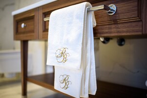 Toalha branca com iniciais bordadas pendurada em um porta-toalhas de madeira em um banheiro.