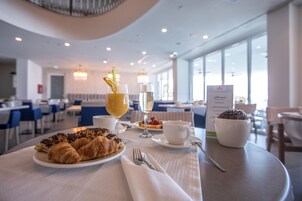 Mesa de jantar com croissants, um copo de vinho, uma xícara de café e uma saladeira numa sala de jantar clara e moderna, com janelas amplas e cadeiras azuis.