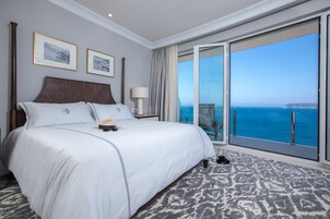 Quarto de hotel com uma cama grande, uma mesinha de cabeceira, uma luminária e vista para o oceano através de uma porta de correr de vidro.