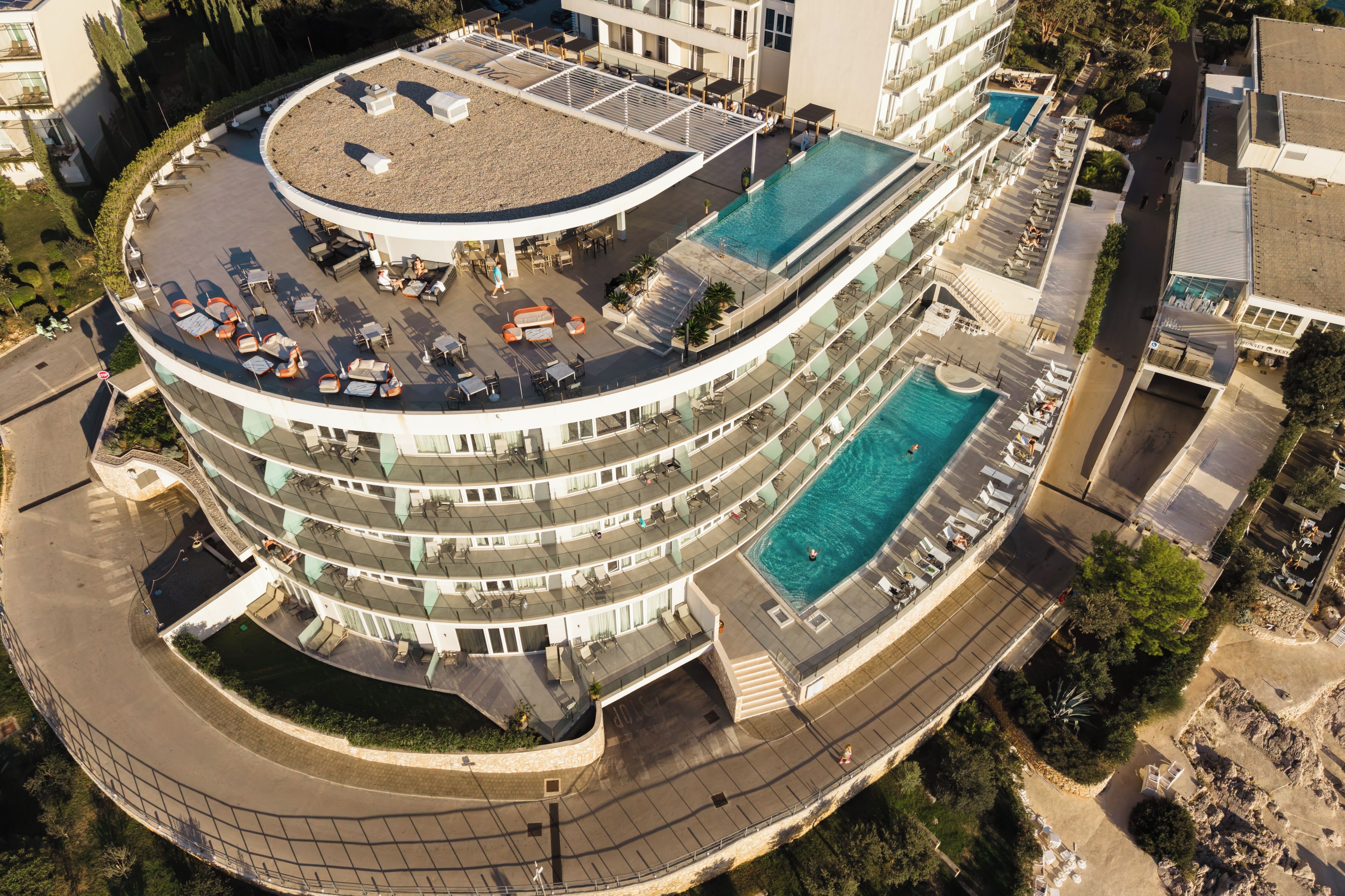 Un hotel de varios niveles con piscina en la azotea, mobiliario exterior y un camino de acceso circular.