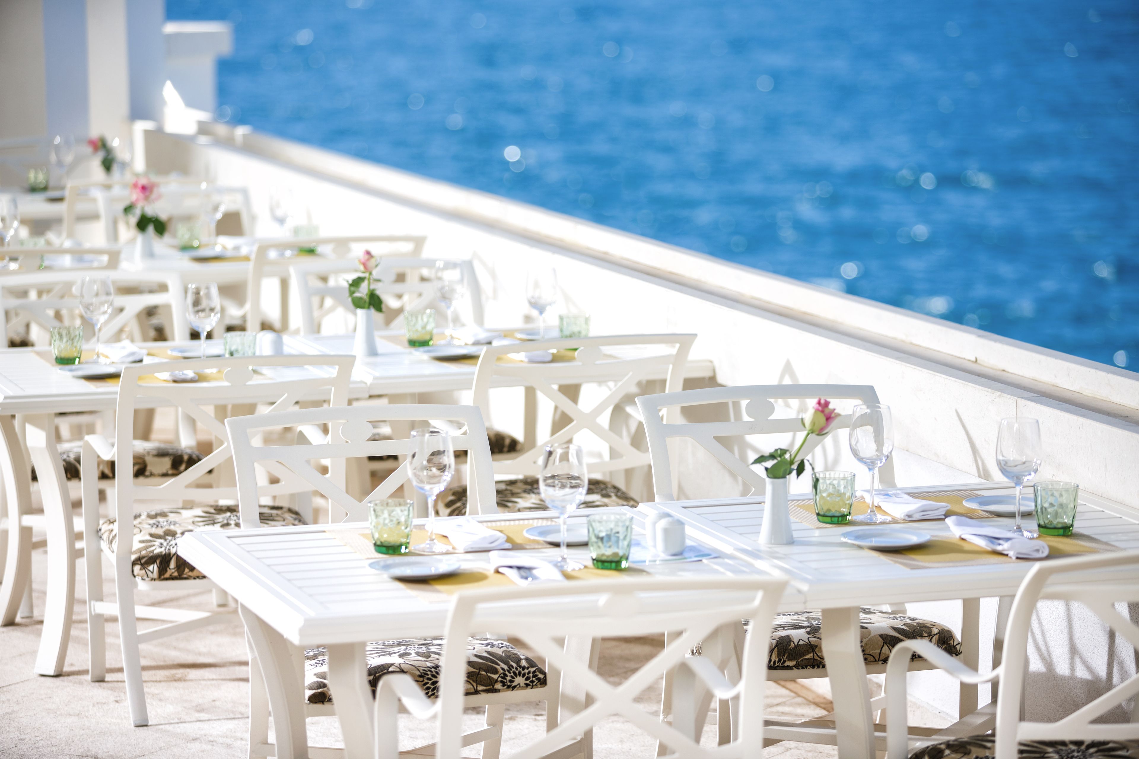Ein Restaurant am Meer mit weißen Tischen und Stühlen, jeweils gedeckt mit Tellern, Gläsern und Besteck.